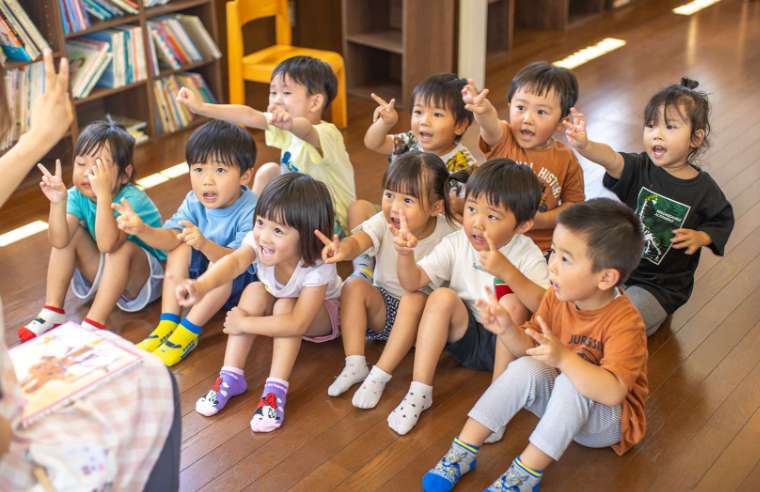 園児達が床に座って先生と同じ手のポーズをしている写真